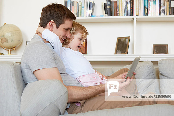 Weibliches Kleinkind sitzt mit seinem Vater auf dem Sofa und schaut auf ein digitales Tablett