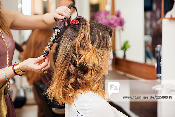 Friseur mit Lockenstab am langen braunen Haar des Kunden im Salon