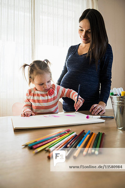 Weibliches Kleinkind am Tisch mit schwangerer Mutter zeichnet im Skizzenbuch