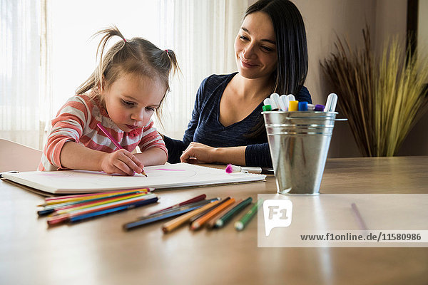 Weibliches Kleinkind am Tisch mit Mutter  die im Skizzenbuch zeichnet