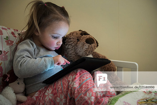 Weibliches Kleinkind sitzt aufrecht im Bett und benutzt Touchscreen auf digitalem Tablett