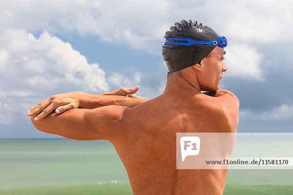 Rear view of muscular male swimmer on beach warming up