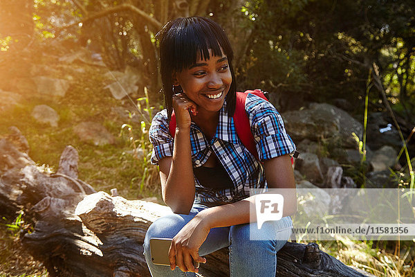 Junge Frau im Wald  auf umgestürztem Baum sitzend  Smartphone in der Hand  Kapstadt  Südafrika