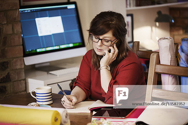Eine Frau sitzt mit einem Smartphone und macht Notizen mit einem Bleistift. Computerbildschirm und Tastatur. Ein mit Stoff und Bastelmaterial gedeckter Tisch.