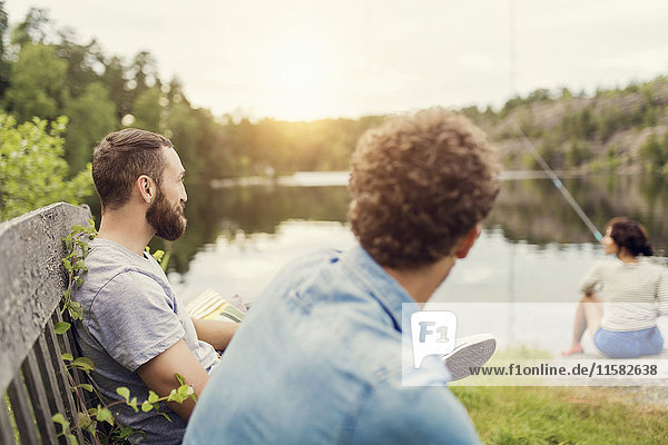 Men sitting on wooden bench while female friend fishing at lakeshore