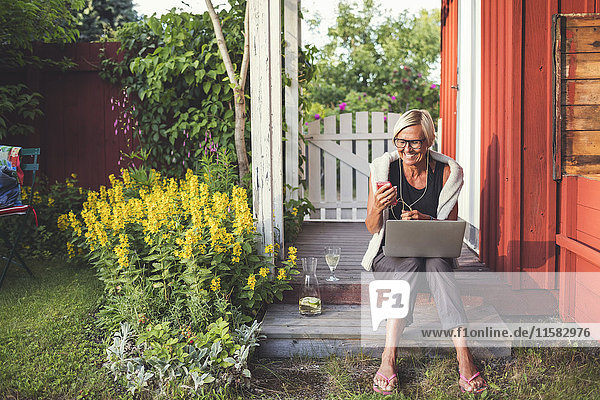 Fröhliche Frau mit Handy und Laptop  die an einem sonnigen Tag im Garten sitzt.