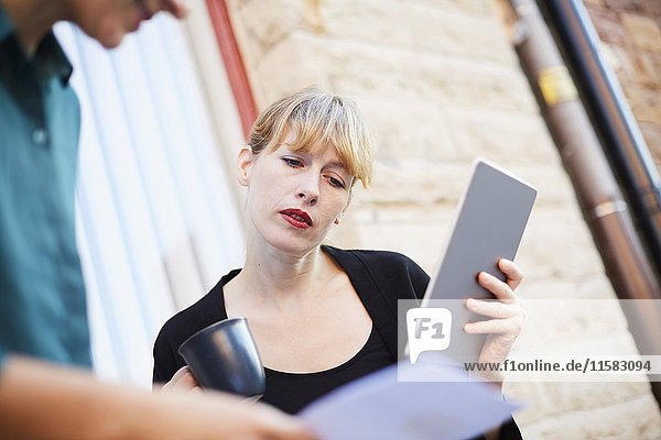 Mid adult businesswoman holding digital tablet and coffee cup while discussing with colleague outside office