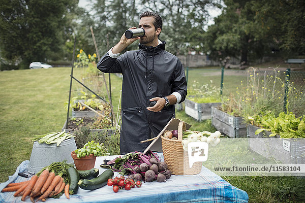 Mid adult man drinking coffee while standing by freshly harvested vegetables on table at urban garden
