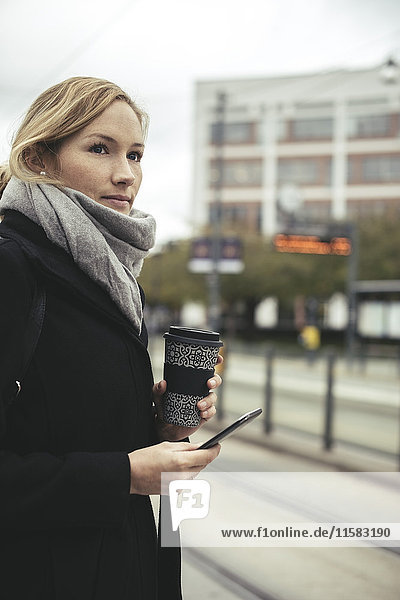 Geschäftsfrau mit Smartphone und Einweg-Kaffeetasse an der Straßenbahnhaltestelle