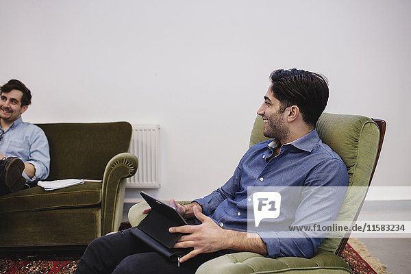 Junger Mann  der auf einem Sessel im Kreativbüro sitzt und einen Kollegen ansieht.