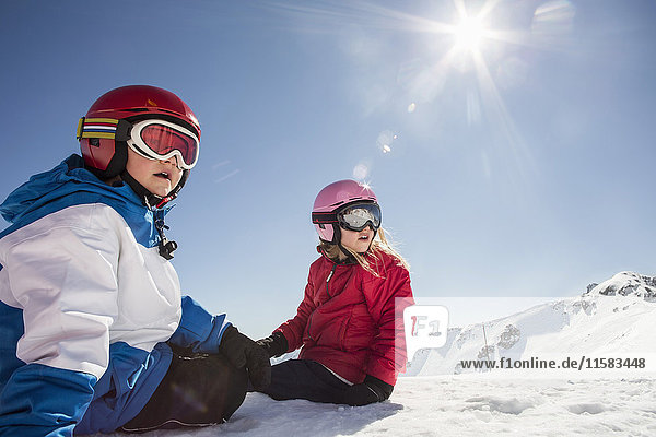 Surprised siblings sitting on snowcapped mountain against clear sky