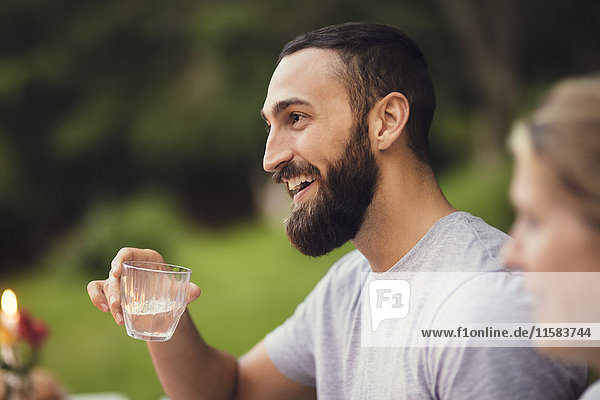 Lächelnder Mann hält Trinkglas  während er mit einem Freund auf einer Gartenparty sitzt.