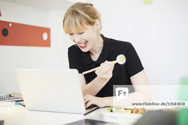 Lächelnde Geschäftsfrau beim Mittagessen mit dem Laptop im Büro