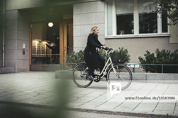 Full length of mid adult businesswoman riding bicycle by city building