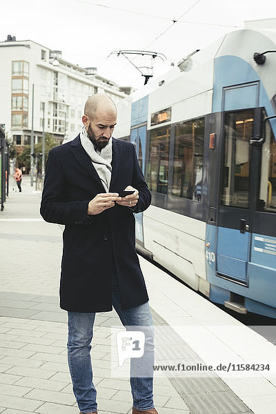 Mittlerer Erwachsener Geschäftsmann mit Handy auf dem Bürgersteig mit der Straßenbahn in der Stadt