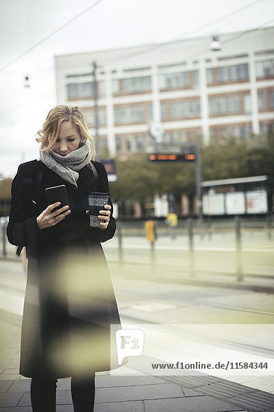 Mittlere erwachsene Geschäftsfrau  die ein Handy benutzt  während sie eine Einweg-Kaffeetasse auf dem Bürgersteig in der Stadt hält.