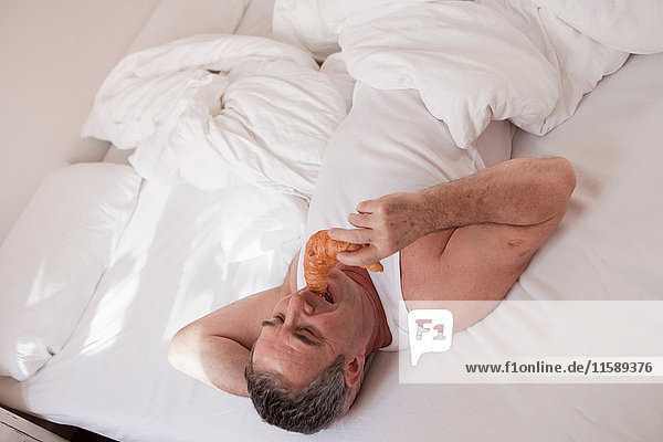 Mann  der auf dem Bett liegt und Croissant isst.