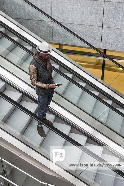 Junger Mann steht auf der Rolltreppe und schaut auf sein Handy.