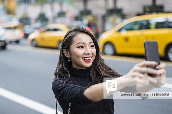 USA  New York City  Manhattan  Porträt einer lächelnden jungen Frau  die sich selbst mit dem Smartphone fotografiert.
