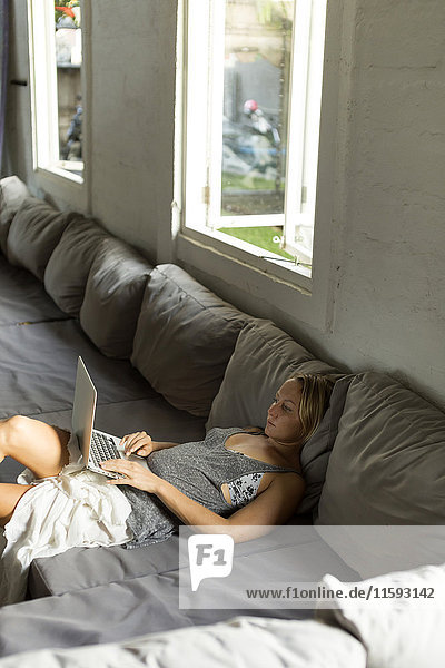 Blonde Frau auf der Couch liegend mit Laptop