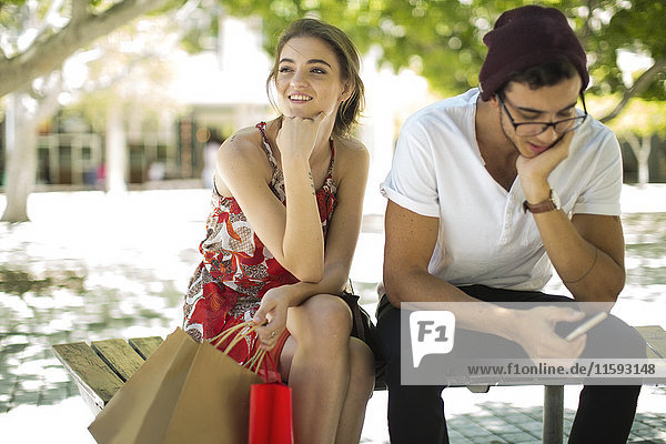 Junges Paar sitzt auf Bank mit Einkaufstaschen und Handy