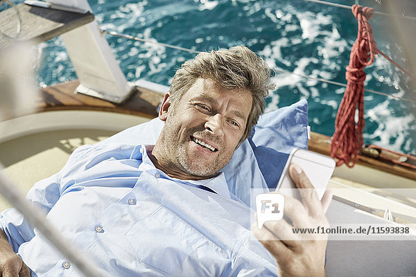 Porträt eines lächelnden  reifen Mannes  der auf dem Deck seines Segelbootes liegt und auf das Handy schaut.