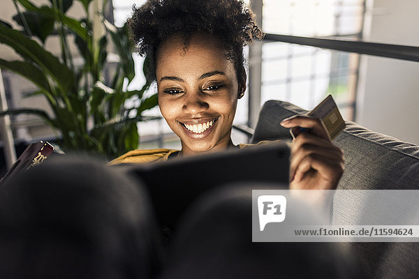 Lächelnde junge Frau auf Couch mit Kreditkarte und Laptop