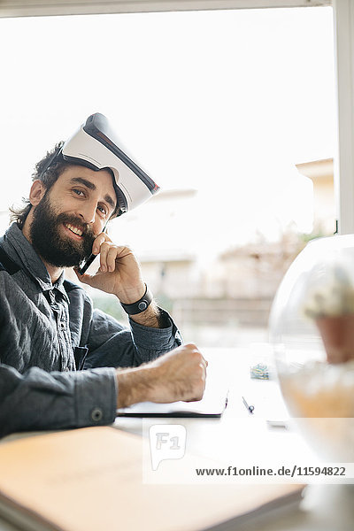 Porträt eines lächelnden Mannes mit Virtual Reality Brille am Telefon in seinem Büro