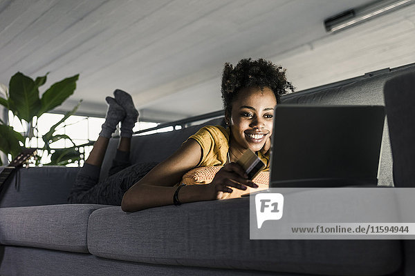 Lächelnde junge Frau auf der Couch liegend mit Kreditkarte und Laptop