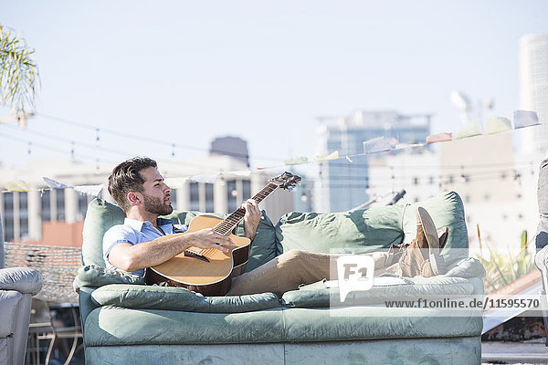 Young man on rooftop lying on sofa and playing guitar