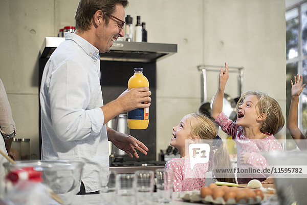Father giving orange juice to excited girls in kitchen