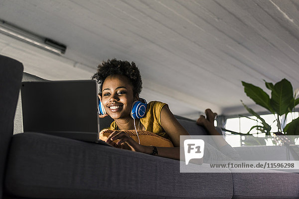 Lächelnde junge Frau mit Kopfhörer auf der Couch liegend mit Laptop
