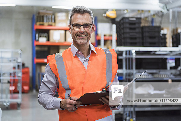 Portrait of smiling man in factory hall wearing safety vest holding clipboard