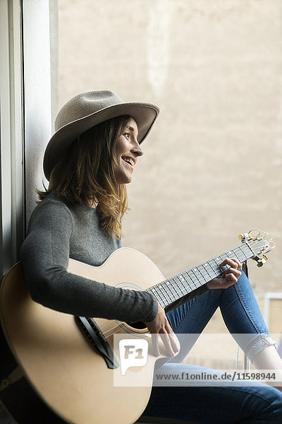 Lächelnde junge Frau sitzt im Fensterrahmen und spielt Gitarre.