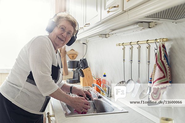 Porträt einer älteren Frau  die beim Spülen Musik mit Kopfhörern hört.