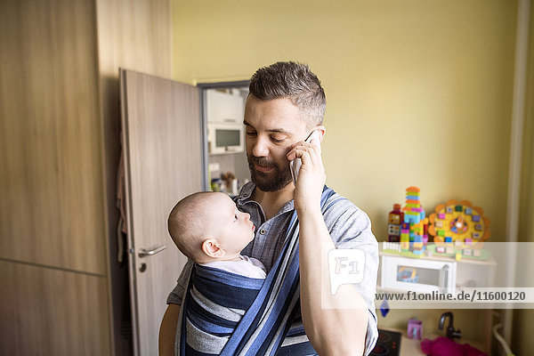 Father with baby son in sling at home talking on cell phone