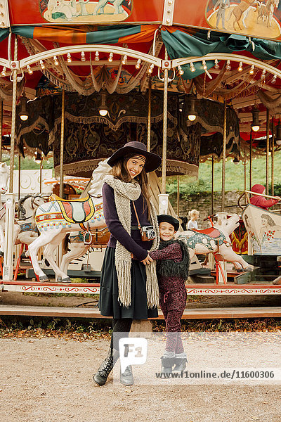 Happy young woman and little girl standing in front of children's carousel at fair