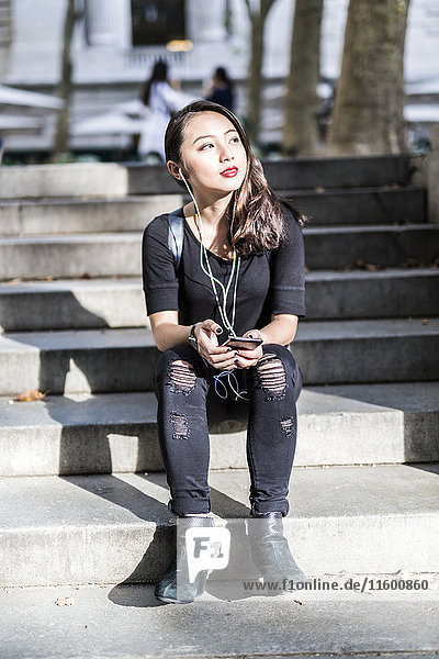 Porträt einer jungen Frau in Schwarz  die auf einer Treppe sitzt und Musik mit Kopfhörer und Handy hört.