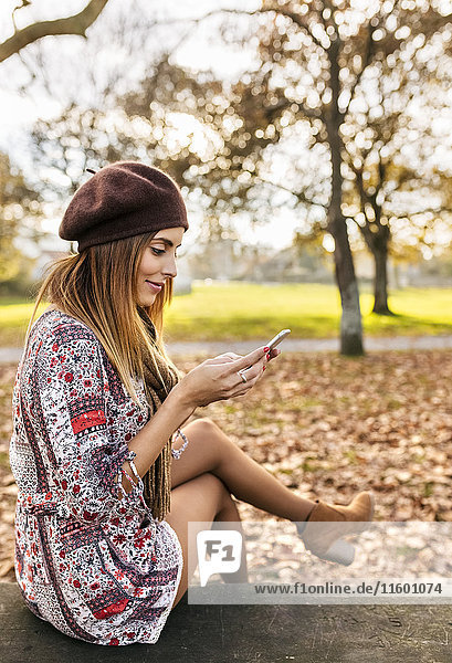 Lächelnde junge Frau sitzt auf der Bank im herbstlichen Park und schaut auf das Handy.