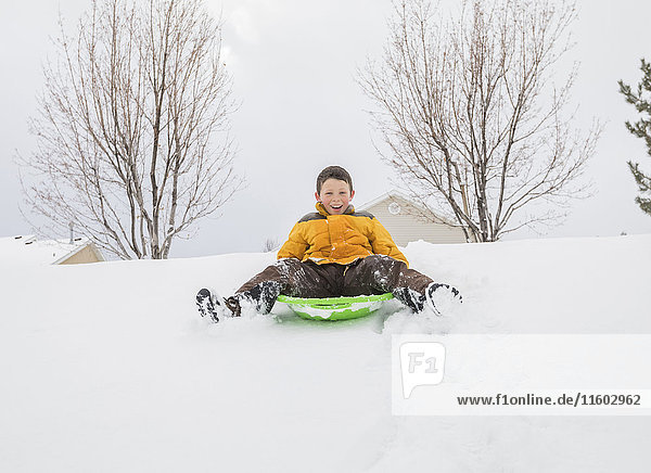 Lächelnder Junge  der auf einem Rodelschlitten auf einem Hügel im Winter rutscht