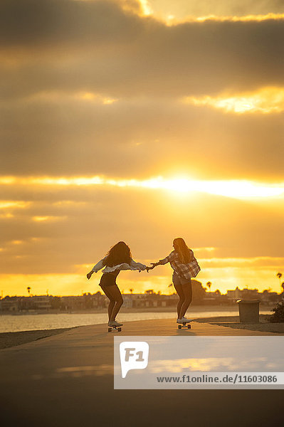 Girl holding hands while skateboarding on waterfront at sunset