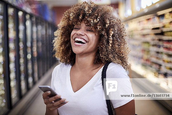 Schwarze Frau hält Handy lachend im Lebensmittelgeschäft