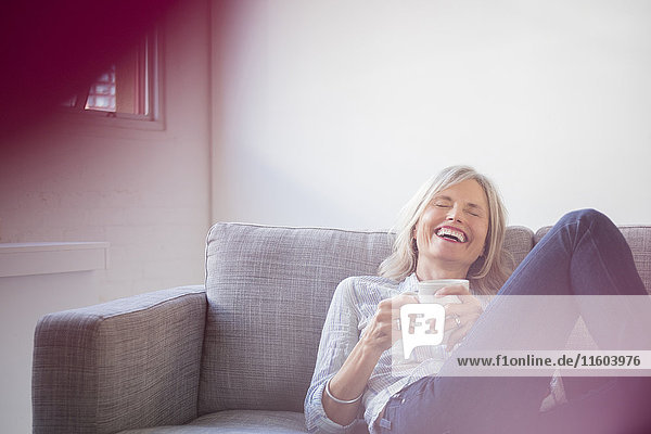 Laughing Caucasian woman sitting on sofa drinking coffee