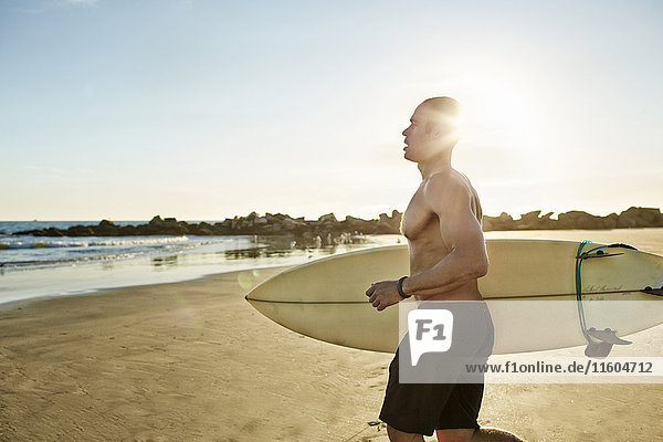 Gemischtrassiger Mann läuft mit Surfbrett am Strand
