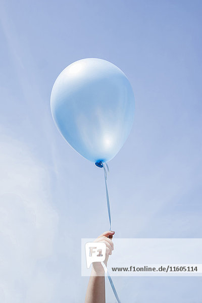 Hand eines kaukasischen Mädchens  das einen blauen Luftballon im blauen Himmel hält