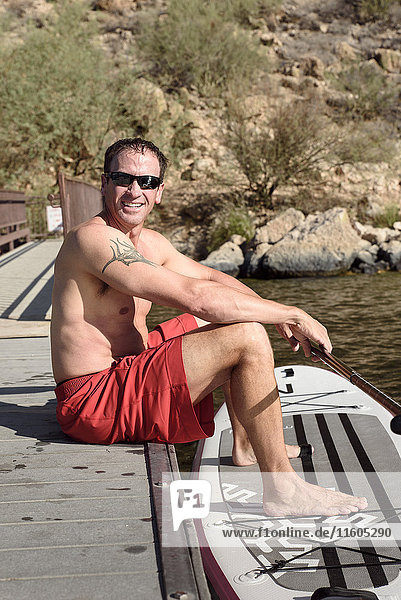 Hispanic Mann sitzt auf Dock mit Paddleboard am Fluss