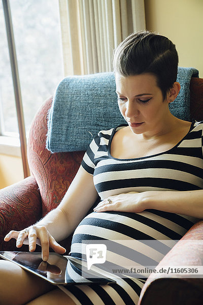 Schwangere kaukasische Frau  die in einem Sessel sitzt und ein digitales Tablet benutzt