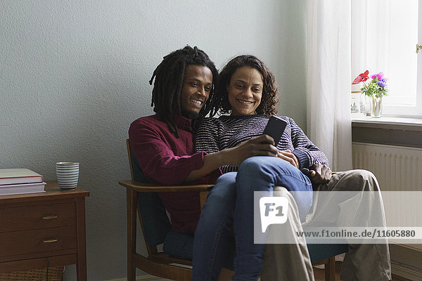 Lächelndes multiethnisches Paar  das zu Hause auf dem Sessel sitzt und ein Smartphone benutzt.