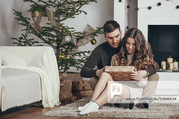 Junges Paar  das zu Weihnachten auf einem Teppich zu Hause sitzt und sich eine Geschenkbox ansieht.