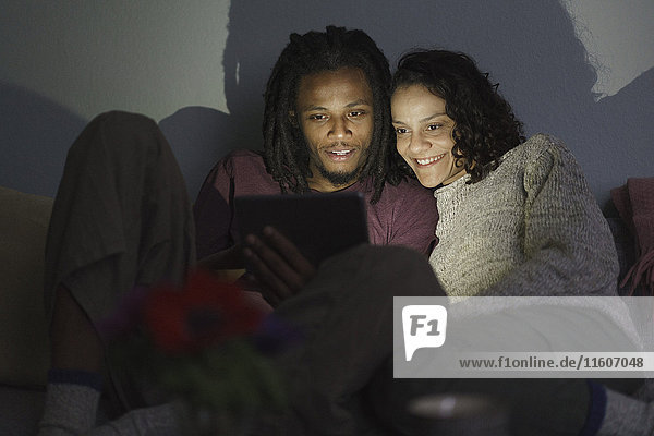 Lächelndes multiethnisches Paar mit digitalem Tablett im dunklen Wohnzimmer